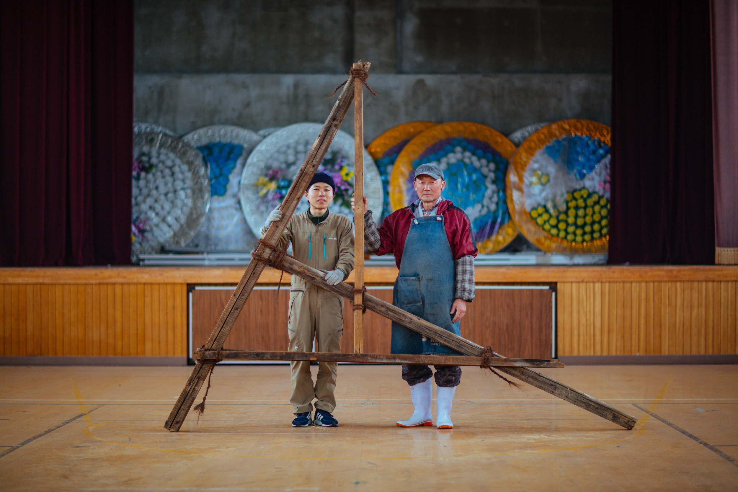 Red Dot School alumni Yosuke Tsurui and board member Yoshihige Kawamoto (Isso) in the Sunoue Elementary School gymnasium, with a 1:1 fragment model of the new structure. The connections are secured with hand-made shuronawa twine from island palm trees.