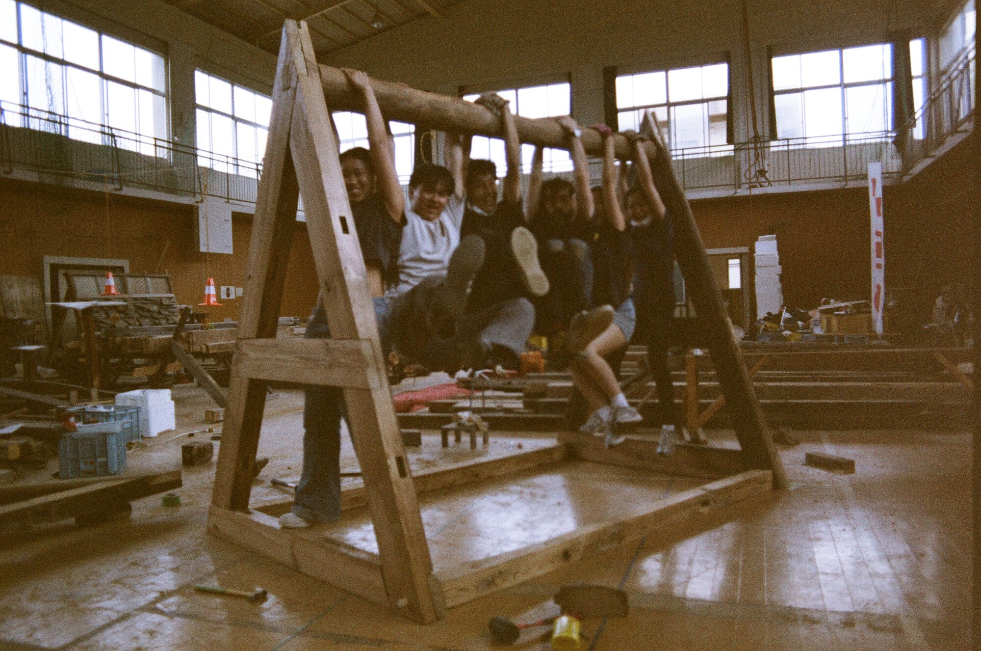 Studio members hanging in a row from the ridge log of a full-size timber A-frame raised on the floor of the old school gymnasium, salvaged lumber and sawhorses laid out around them