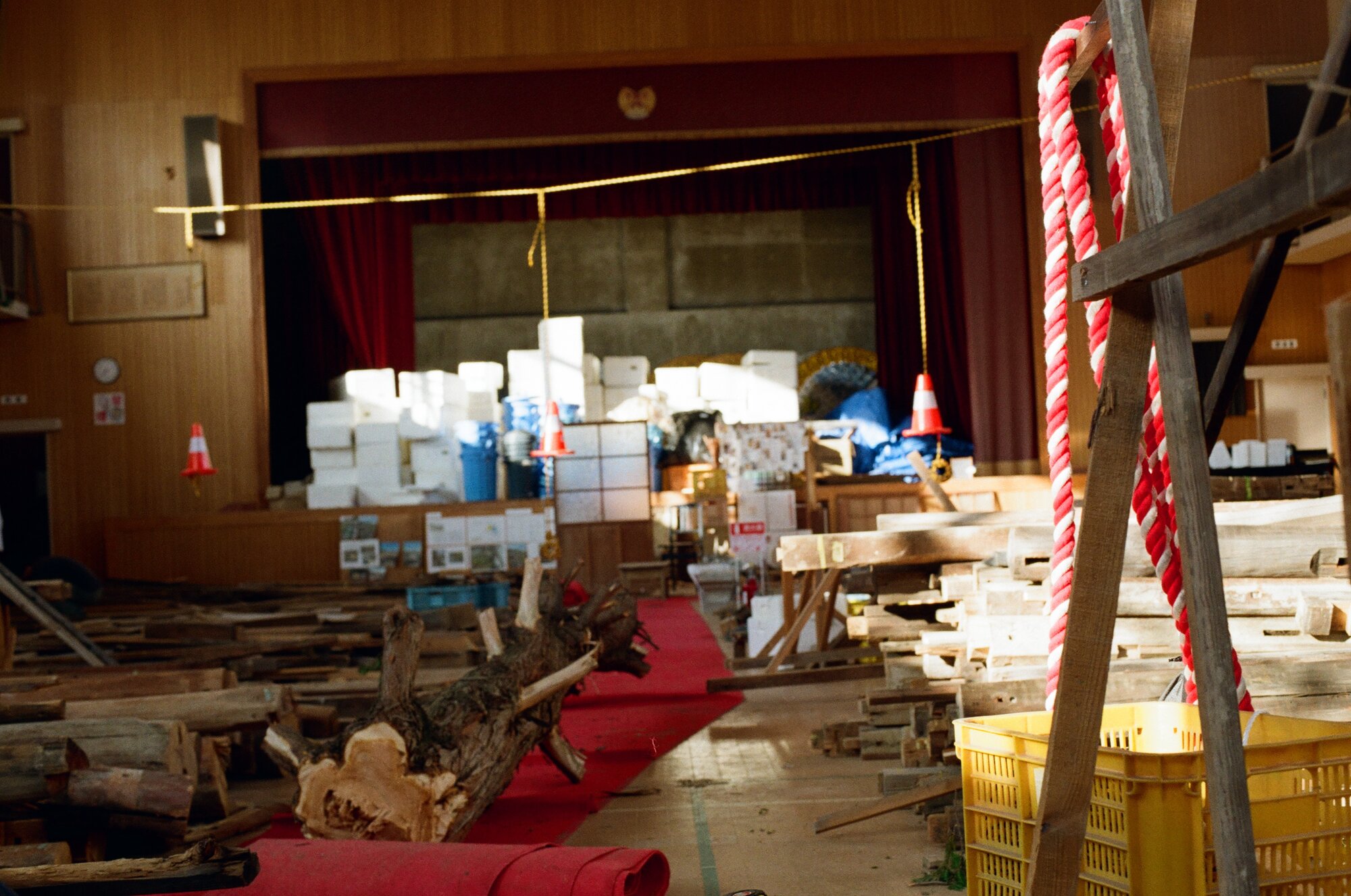 The old school auditorium in use as a salvage yard: recovered timber laid out in sorted rows across the floor, stacked crates filling the curtained stage