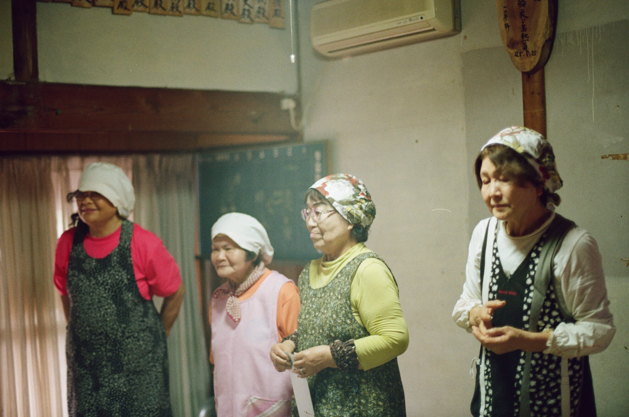 Four island women in work aprons and headscarves standing in a community room, one holding a printed sheet, with a chalkboard and rows of wooden donor plaques behind them