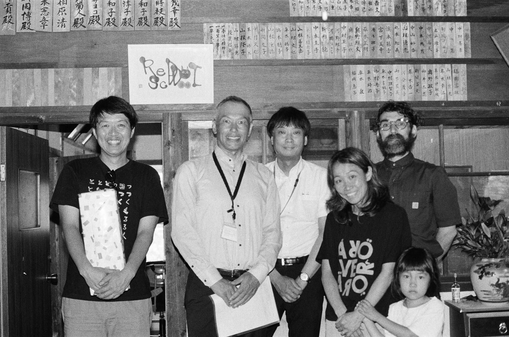 Five adults and a child standing together in a traditional timber-framed room, one holding a wrapped gift, beneath rows of wooden dedication plaques, with a hand-lettered Red Dot School sign on the post behind them
