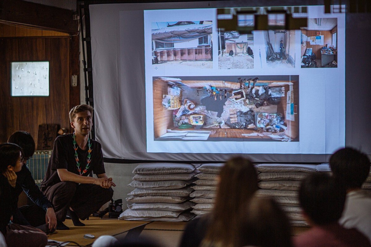 A lecture inside the Kakumeisha classroom: students seated on tatami around a stack of zabuton cushions, listening to a faculty member