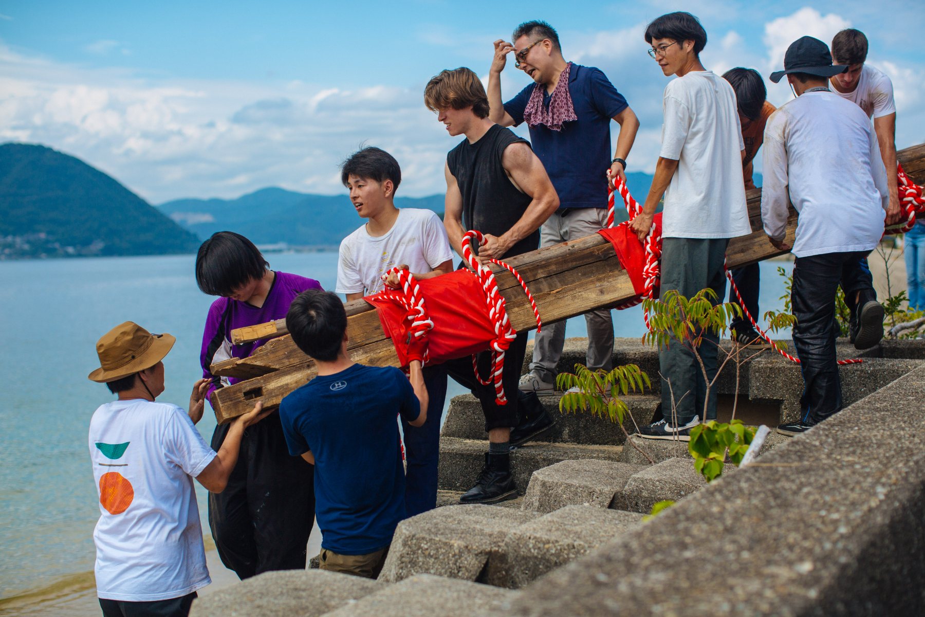 Students, faculty, and Sagishima community members shoulder a bundle of salvaged timber together up the seawall, the bundle wrapped in red cloth and red-and-white festival rope