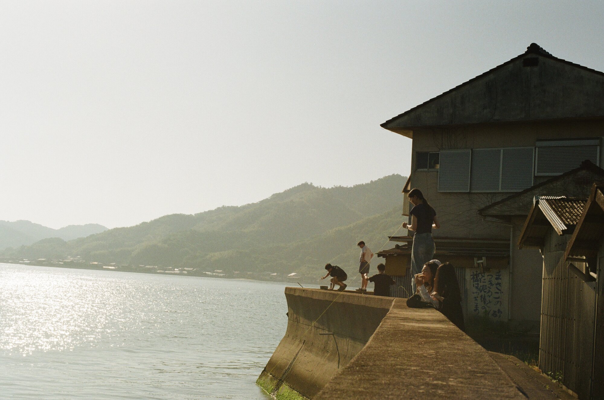 Studio members gathered along the concrete seawall at Sunoue beside a weathered island house, some fishing with hand lines, some sitting on the wall, the Seto Inland Sea and far hills hazy behind them